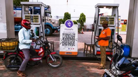 NurPhoto via Getty Images A rider sitting on his moped waits next to a large PhonePe placard at a petrol pump in India.