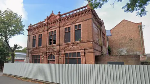 Google A two-storey building behind a fence with part of the pavement, which is in front of the fence, visible in the bottom left of the photo. Trees and another structure are on the far left of the image.