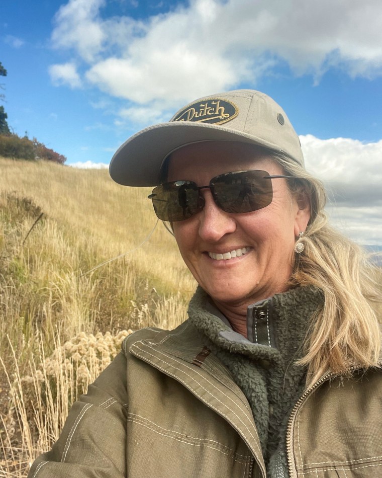 A selfie of Angela Newman smiling while standing outside in a field, wearing a brown coat, sunglasses, and a Von Dutch baseball cap