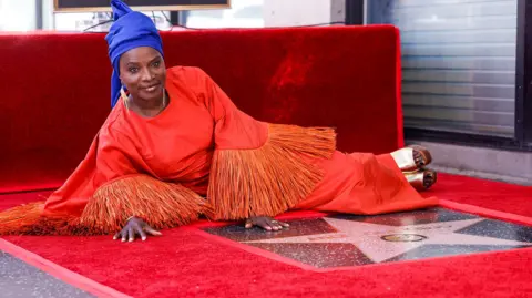 EPA Angélique Kidjo lying down on a red carpet behind her star which is on the ground. She is wearing a red, long-sleeved dress with tasselled sleeves and a royal blue headwrap
