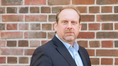 LGIU Carr-West, with greying red hair looks into the camera. He is pictured as a head and shoulders shot, wearing a blue suit and a sky blue shirt. In the background is a brick wall.