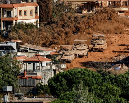 Three Israeli tanks go down a dusty road a village