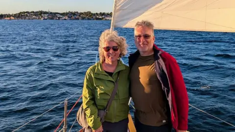 Supplied by Kent Truscott A couple pictured on a boat with blue water behind them and land in the distance. The woman on the right is wearing a green jacket, and the man is wearing a black shirt and a red jacket on top. It is taken during golden hour, with a golden glow from the sun shining on them and the boat.