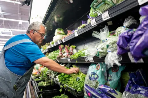 Getty Images A man wearing a blue t-shirt and dungarees picks coriander from a fresh produce shelf in a supermarket.