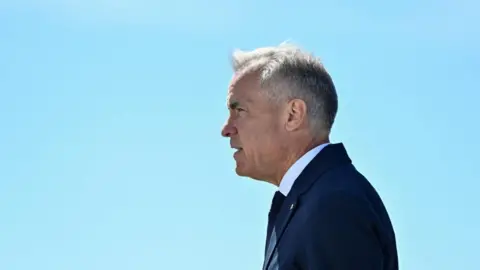 Reuters Mark Carney stands in profile against a clear blue sky. His hair is short and grey, and he wears a navy suit and tie and a white short.