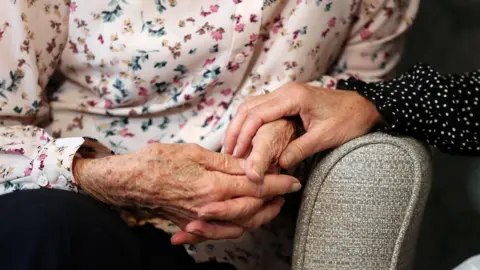 PA Media An elderly woman sitting down on a chair. She is wearing a light pink blouse with a pink and blue floral design across it. Her hands are clutched together with another woman who is wearing a black and white polka dot blouse holding one of her hands.