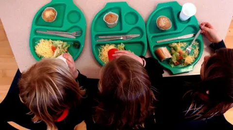 PA Media Three girls sitting at a table with three green school meal trays with cutlery, cheese salad, and a muffin on them. The view is overhead and two are drinking from cups.
