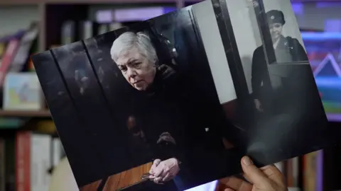A hand holds a printed out photograph of Nadezhda Buyanova, a 68-year-old Ukrainian-born paediatrician in the dock at court, guarded.
