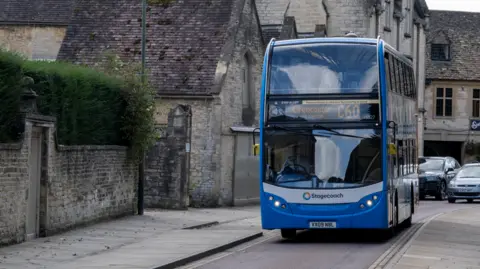 Getty Images A blue bus with 'Cirencester' as the destination on it drives through a single laned road next to a quaint looking brick wall.