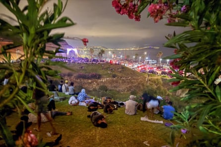 People sitting on a grassy hill at night looking out to sea where lights shine