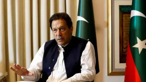 Reuters Former Pakistani Prime Minister Imran Khan speaks with Reuters during an interview, in Lahore, Pakistan March 17, 2023. He can be seen seated with his hand outstretched and speaking. Two Pakistani flags are behind him