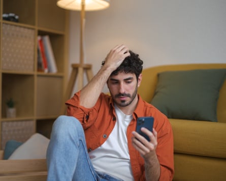 A bearded young man sitting on the floor of his living room looking slightly distressed as he looks at his mobile phone.
