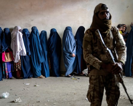 Afghan women wearing burqas stand in a line beside a soldier in uniform holding a rifle.