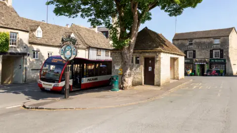 Getty Images A red bus drives through a picturesque village with a bin and small building with a brown door next to it.