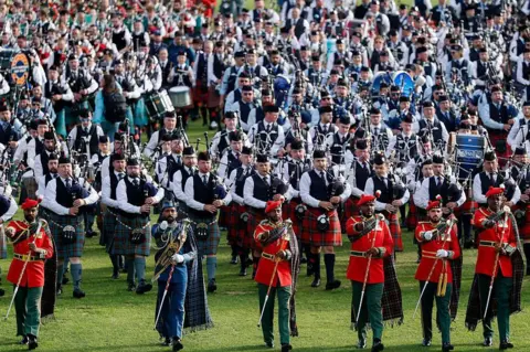 Getty Images A large gathering of drummers and pipers parading on a field. They are all in traditional outfits and are being led by marching leaders.