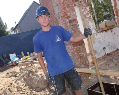 A young man named Kobe wears a hard hat and gloves at a construction site holding a tool