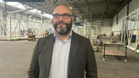 Michael has a full beard, black with patches of grey hair around his chin. Suited and wearing dark rimmed spectacles, he is standing in an air hanger.