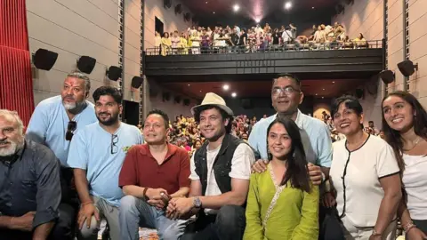 Kapil Rijal A group of people crouch on a stage with an auditorium full of people behind them.