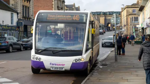 Getty Images A white bus pulls up at a bus stop as people walk by