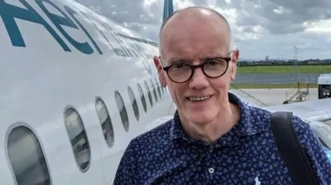 John Bittles A man, John Bittles pictured beside an aeroplane. he is wearing a navy shirt, a black bag strap on his right shoulder and black glasses. He is bald.
