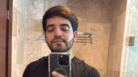 Instagram/César Gastélum César Gastélum looks into a mirror as he takes a selfie. He has a beard and black hair. He is wearing a black T-shirt and a gold chain around his neck