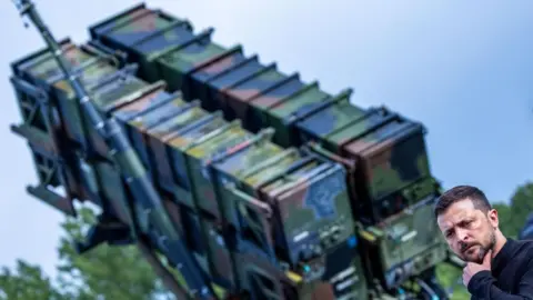 EPA-EFE/REX/Shutterstock Ukraine's President Volodymyr Zelensky stands in front of a 'Patriot' anti-aircraft missile system during a 2024 visit to a military training area in Germany. He is dressed in black and has one hand on his chin.