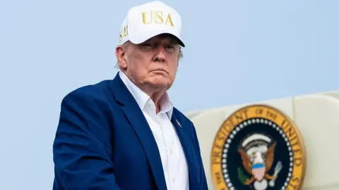 Getty Images Trump, wearing a suit and no tie and a white "USA" hat, descending from airplane steps