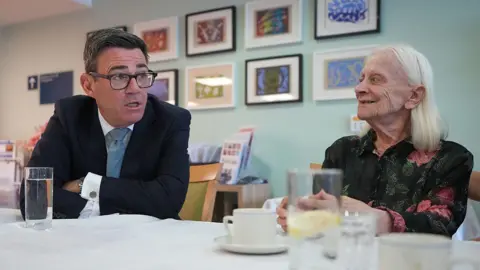 Getty Images Andy Burnham sits at a table in a care home, talking face-to-face with a resident. The prime minister is in a dark suit and glasses and leans forward while speaking, and the other, with long white hair, sits opposite. Several glasses of water and cups are on the table, with framed artwork displayed on the wall behind them.
