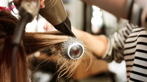 Getty Images A hair-styling treatment is being carried out in a salon. The stylist's hands are partially visible holding the dryer and brush as the hair is pulled taut, a common technique used to smooth and straighten hair during a blow-dry.