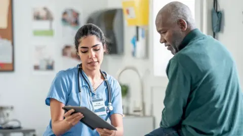 Getty Images/FatCamera A nurse in blue scrubs with a stethoscope consults with a Black patient in a clinic, using a tablet. Warm, professional healthcare scene highlighting patient care and medical teamwork.