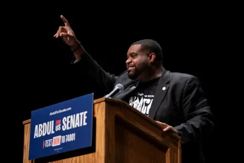 Getty Images Donavan McKinney wearing a T-shirt and dark suit speaks from a podium at The People v The Powerful rally in Detroit in July