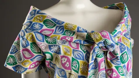 The Harris Close-up of the shoulders of a cream mannequin, wearing a cotton Bardot-necked dress tied at the front. The pattern has decorative leaf motifs in blue, green, yellow and pink on a white background.