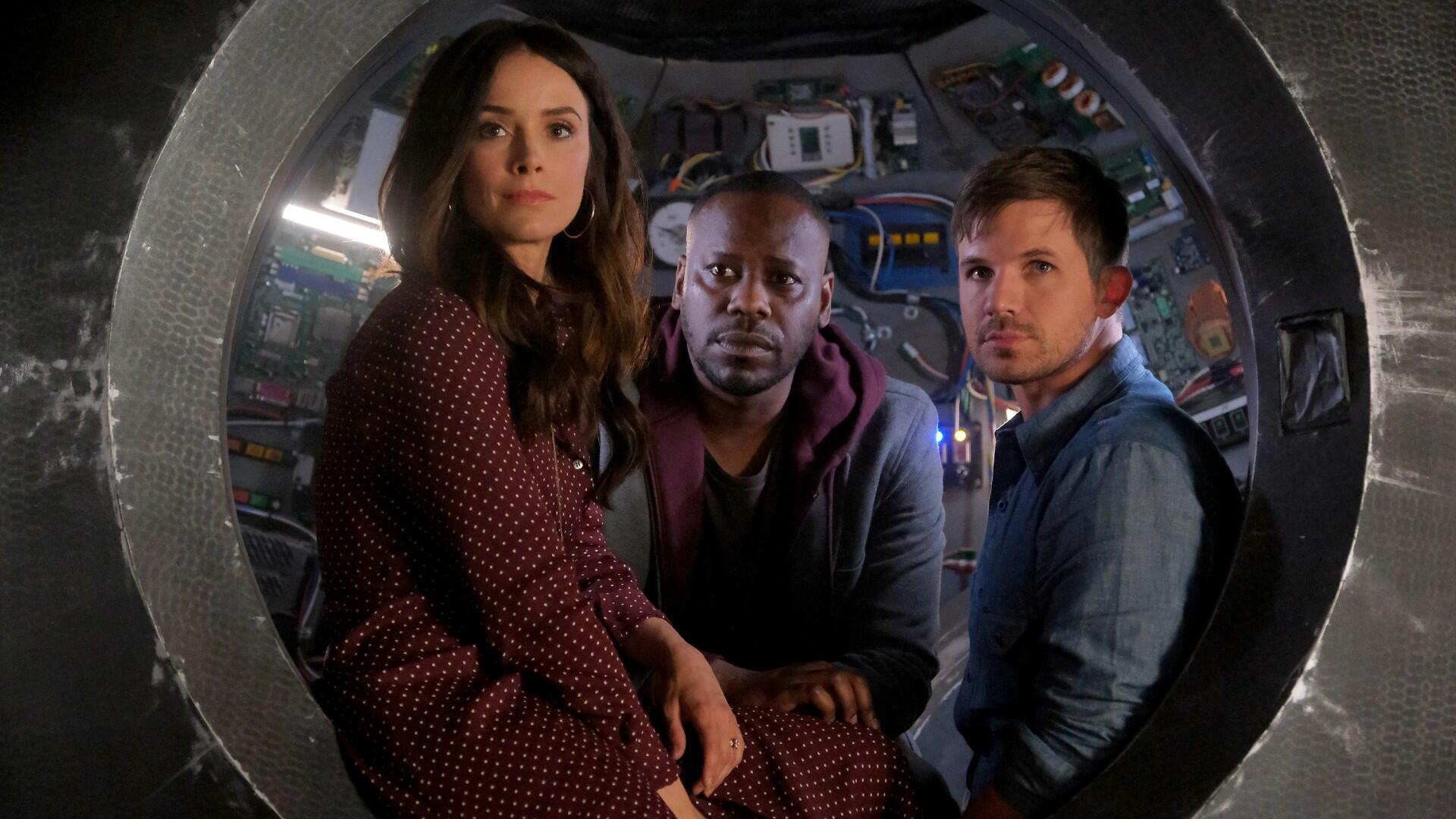 A white woman, a Black man and a white man sit in the cockpit of a time machine.