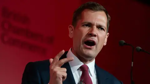 Anadolu via Getty Images Robert Jenrick makes a hand expression as he speaks at a podium in front of a red-lit background.