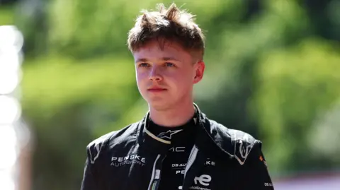 Getty Images/Formula E Taylor Barnard wears a black racing suit. He is taking an earpiece out of his ear and looking off into the distance.