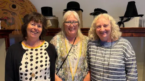BBC A woman in a black cardigan and spotted shirt, another woman with a colourful yellow top and glasses, and another woman with a blue, stripy shirt, stand in front of a hat collection.