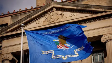 Getty Images Close up of a DOJ flag (a rectangular base background of ultramarine blue, bearing an eagle on a shield, a scroll and the inscription 'Department of Justice.' The eagle faces to the left, with its left claw holding 13 arrows with the tips facing down. Its right claw holds an olive branch. The shield consists of a white base, a blue chief and six scarlet stripes. The scroll shall read in bold blue letters, 'QUI PRO DOMINA JUSTITIA SEQUITUR' - meaning, Who for Lady Justice prosecutes) flying in front of the DOJ headquarters