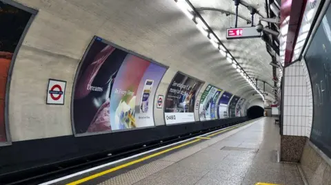 Getty Images An empty platform at Chancery Lane Underground station in London, looking down the curved track tunnel. Large advertising billboards line the tiled wall opposite the platform, illuminated by overhead strip lighting. A dark tunnel entrance is visible in the background, with a London Underground roundel sign mounted on the left wall.