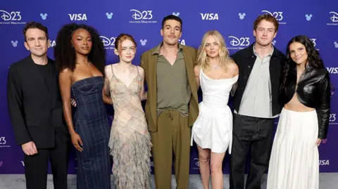 Getty Images The cast of the new X-Men movie all standing up posing for a picture. Jake is in all black, Maya is in a dark blue dress, Sadie is wearing a dress with a rectangle pattern on it, Christopher is wearing an olive blazer and trousers, Samantha is wearing a white dress, Kit has a black jacket on with a stripy shirt on underneath, Inde is wearing a black leather jacket with a white skirt.