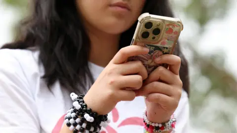 Reuters A teenage girl wearing coloured bracelets sits outside looking down at her iPhone
