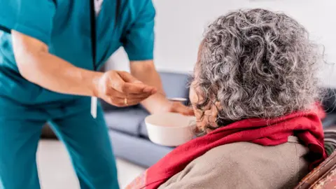 Getty Images A standing, uniformed care worker feeds a seated, elderly woman from a bowl with a spoon