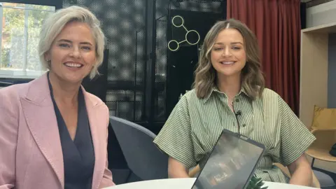 Oonagh and Marie-Clare sit beside each other, a foot or two apart, behind a circular white desk. One has blonde hair and wears a white blazer. The other Has brown hair and wears a green and white striped dress.