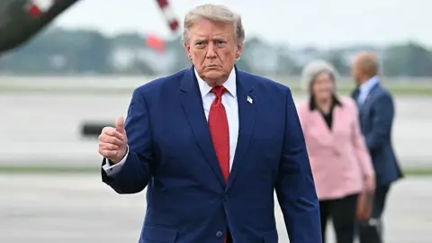 Getty Images US Donald Trump looking serious, giving a thumbs up to the camera. He is wearing a blue suit with a red tie.