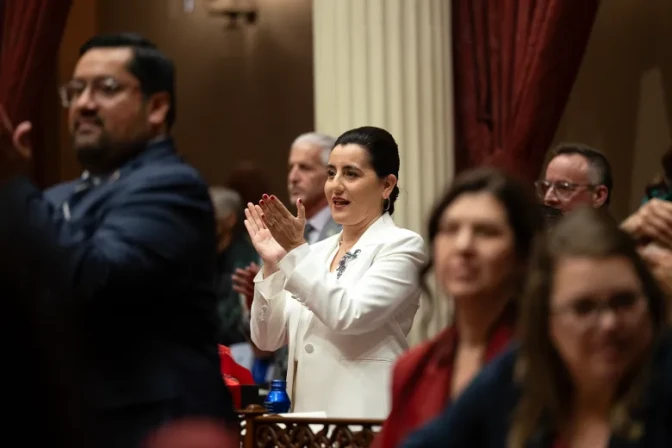 Senate President Pro Tem Monique Limón, a woman with light skin tone, wearing a cream-colored suit, claps her hands as she stands while others around her do the same.