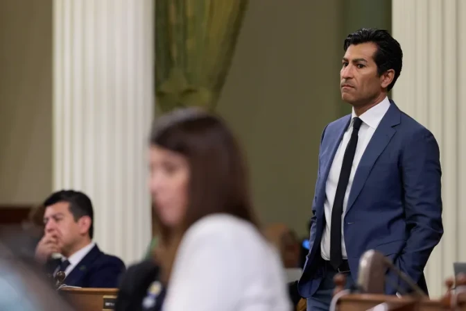 Assembly Speaker Robert Rivas, a man with medium skin tone, wearing a blue suit and tie, stands as he listens to someone out of frame. There are other people in the foreground and background who are out of focus.
