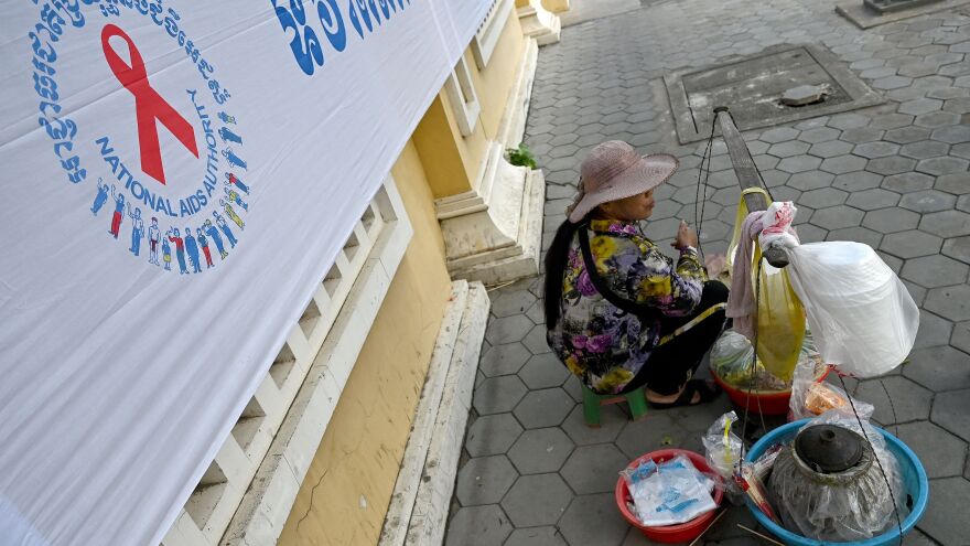 A vendor sits next to a logo for World AIDS Day, along a sidewalk in Phnom Penh on December 1, 2022. (Photo by TANG CHHIN Sothy / AFP) (Photo by TANG CHHIN SOTHY/AFP via Getty Images)