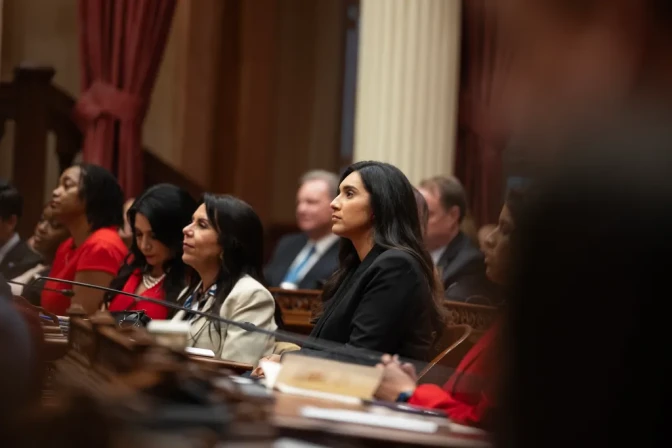 State Sen. Sabrina Cervantes, a woman with medium skin tone, wearing a black coat, sits and listens at a wooden desk along with people doing the same thing.