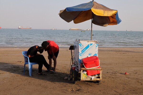 People stand next to a vendor's cart on the beach as cargo ships and offshore service vessels are anchored in the Strait of Hormuz, off Bandar Abbas, Iran, Sunday, May 31, 2026. (Amirhosein Khorgooi/ISNA via AP)
