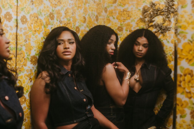 Three woman stand beside each other in front of a mirror. Wall paper decorated with yellow flowers is behind them as each look at different directions in the frame.