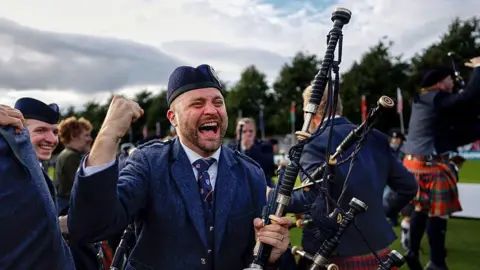 Getty Images A piper from the Simon Fraser University pipe band celebrating their win. His mouth is open in a wide smile and he is punching the air. He is wearing a traditional bagpiper outfit with hat and is holding his bagpipes. There are other pipers behind him.